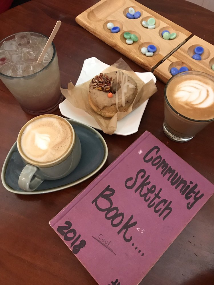 Lattes, a pastry and a mancala board beside the 2018 Community Sketch Book