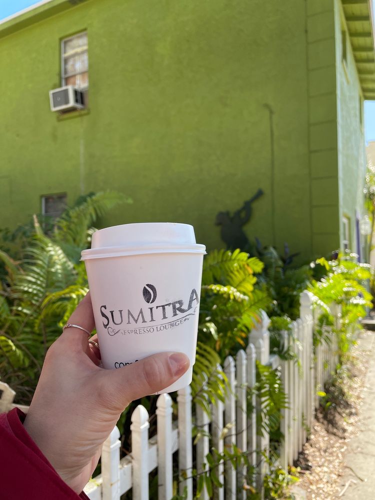 A SumitrA to-go cup held up against a green wall and a white picket fence