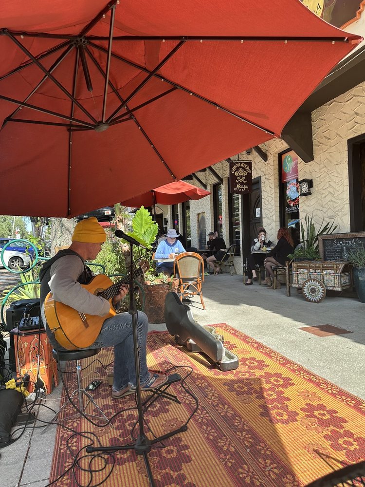 A guitarist playing on a patterned rug under a red umbrella on SumitrA's sidewalk